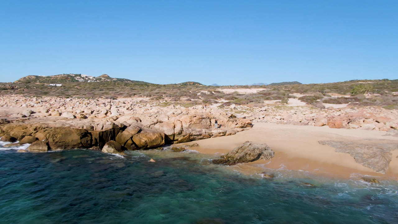 Second frame of Laguna Beach Los Cabos with turquoise water below, darker rock shelves near the beach, pale sand behind them, and no clearly visible people.
