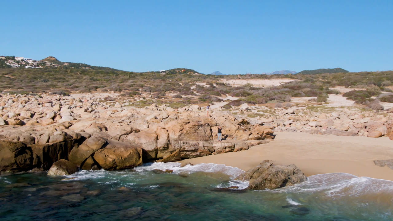 Rocky beach with waves, sand, scrub hills, and a small distant person.