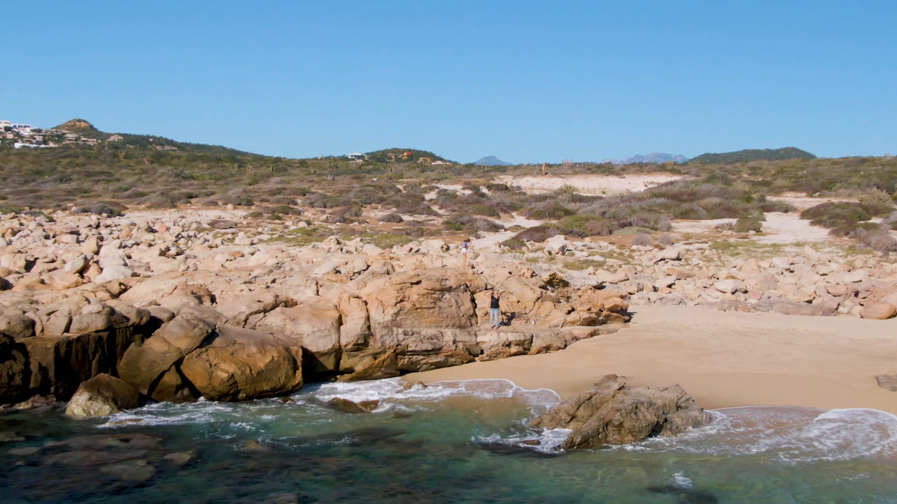 Laguna Beach Los Cabos frame with foamy water reaching the sand, a rounded rock outcrop at the shoreline, dry brush on the dunes, and a distant person beside the rocks.