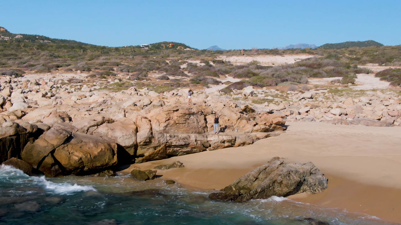 Wide view of Laguna Beach Los Cabos with turquoise water in the foreground, tan rocks and sand at center, scrubby hills beyond, and small people visible on the rocks.