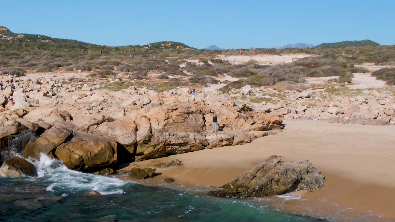 A wave breaks harder against the left-side rocks at Laguna Beach Los Cabos, with open sand, dry brush, hills, and small people still visible in the rocky midground.
