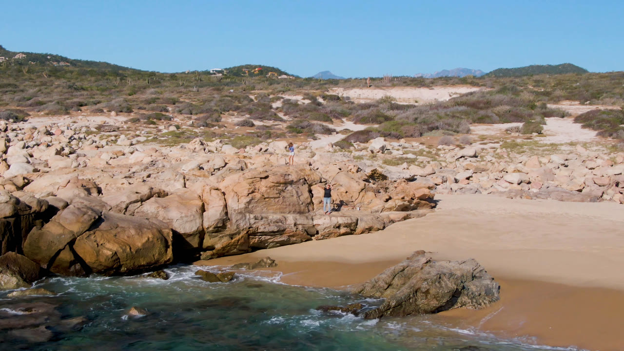 Rocky beach with sand, shallow water, dunes, and blue sky.