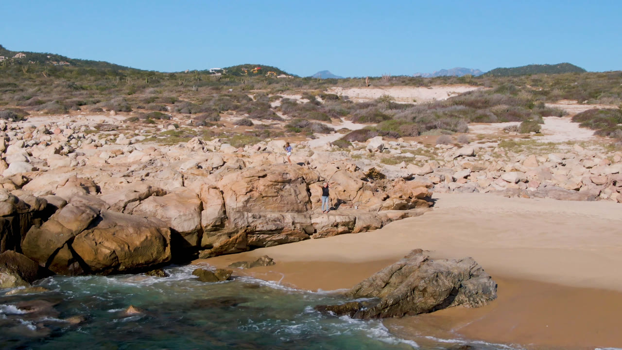 Laguna Beach Los Cabos shoreline with blue-green water at lower left, a central rock outcrop, open tan sand, scrubby dunes, and distant beachgoers near the rocks.