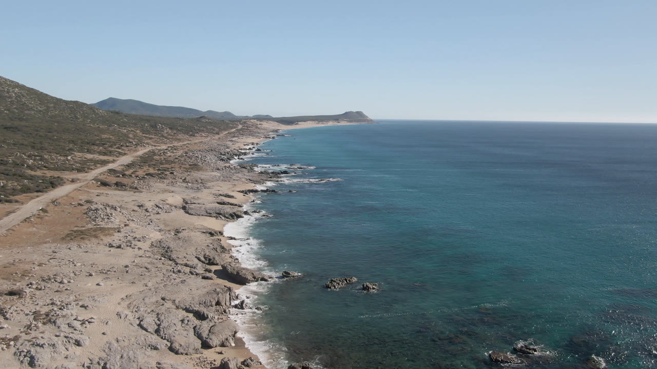 Aerial view of Laguna Beach Los Cabos with tan rocky shoreline, blue ocean, scrub hills, distant headland, clear sky, and no visible people.