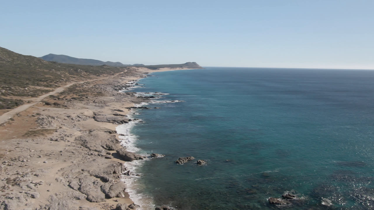 Drone-height view over Laguna Beach Los Cabos showing pale rocks, turquoise shallows, darker offshore water, low hills, a far point of land, and no visible people.