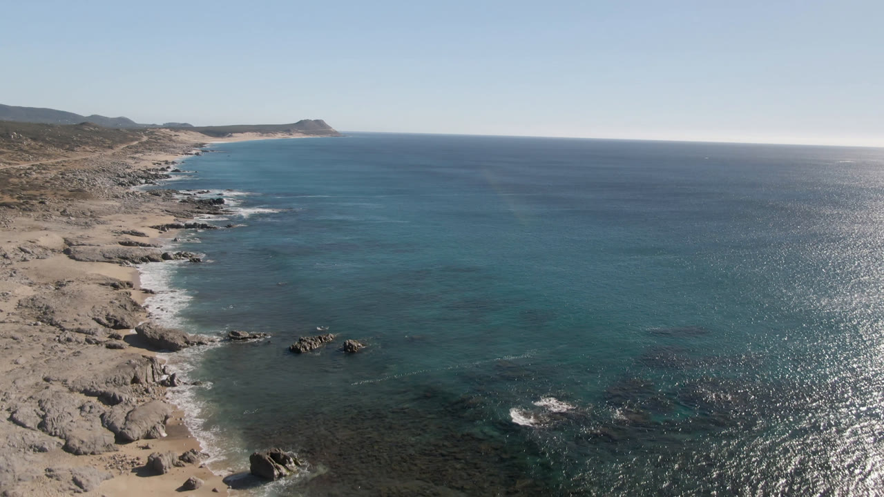 Aerial frame over Laguna Beach Los Cabos showing pale sand, rocky shoreline, blue ocean, a distant headland, and no visible people.