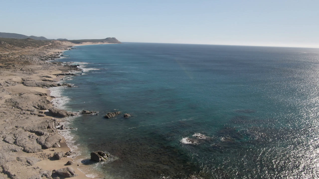 Aerial view at Laguna Beach Los Cabos with the rocky shore extending into the distance, shallow turquoise water near the beach, and no visible people.
