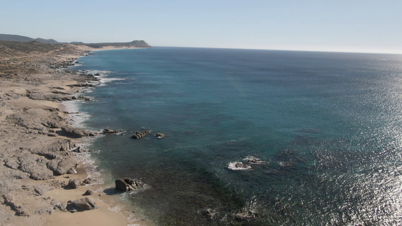 Wide aerial view of rocky coast with sunlit ocean surface.