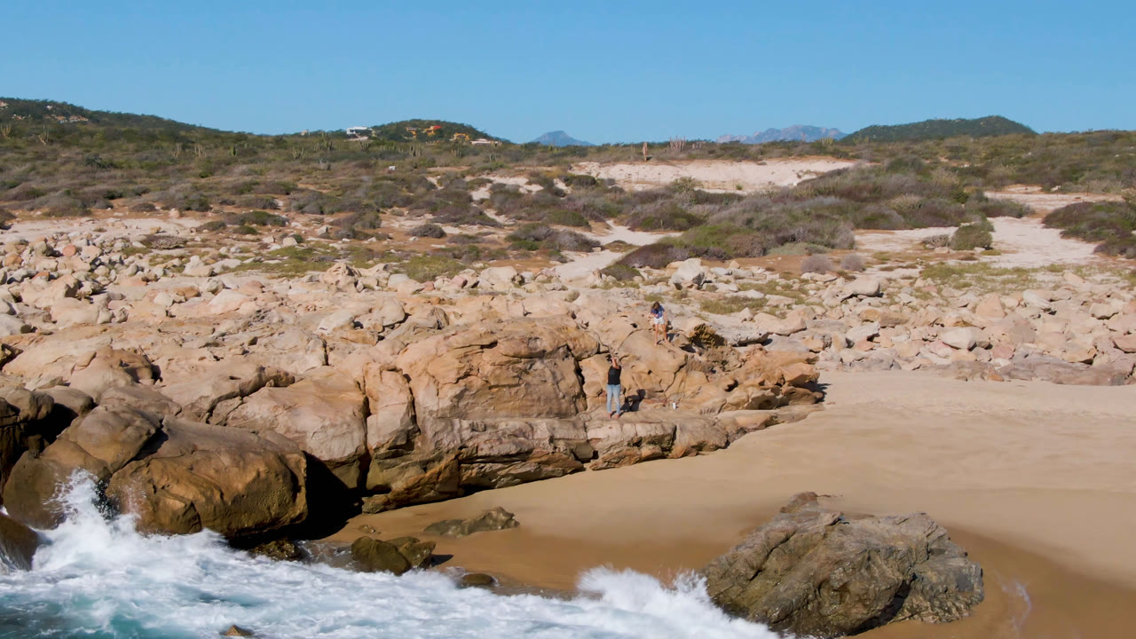 At Laguna Beach in Los Cabos, white water wraps around the lower rocks while the sandy pocket beach, tan boulders, scrubland, hills, blue sky, and small people remain visible.
