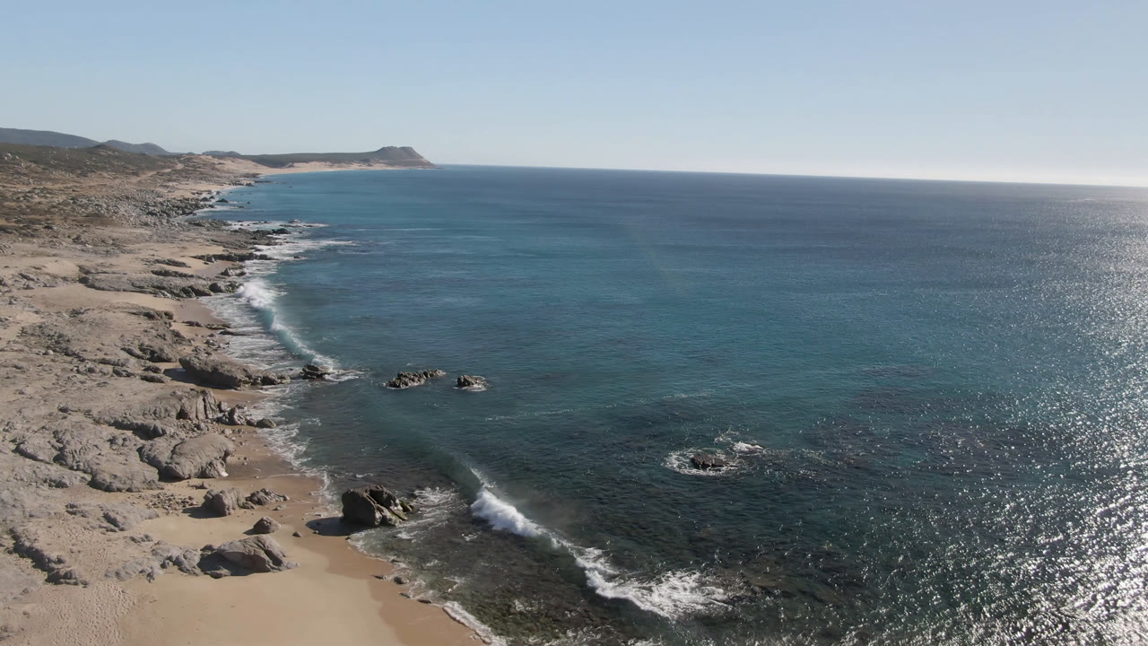 Aerial frame over Laguna Beach Los Cabos showing pale sand, scattered coastal rocks, blue ocean, white surf, distant hills, and no visible people.