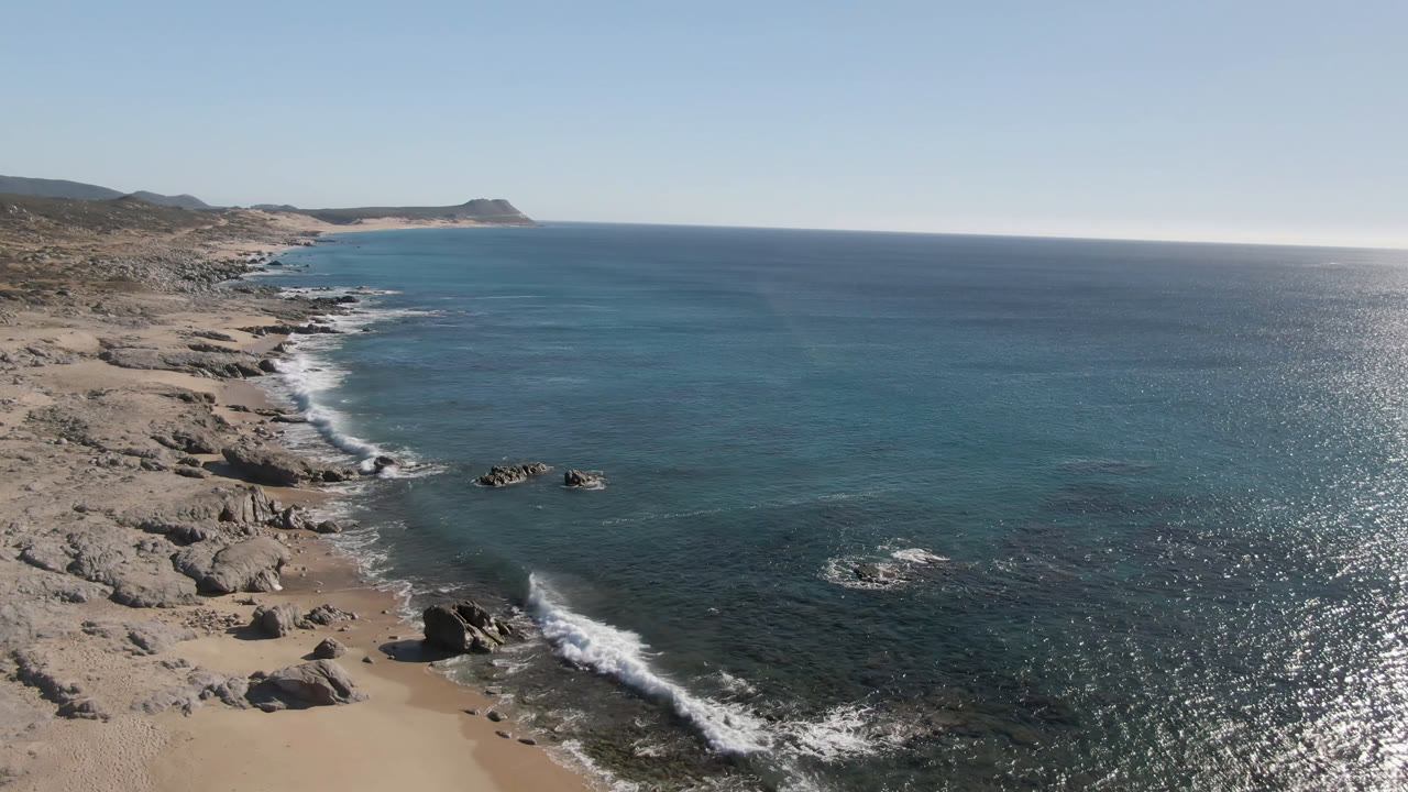 Laguna Beach Los Cabos coastline from above, with surf tracing the beach edge, rock clusters in the shallows, open ocean to the right, and no visible people.