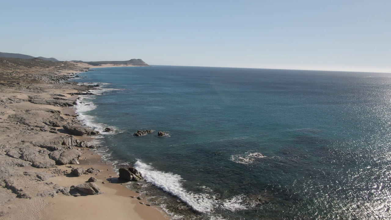 Wide aerial coastline with rocks, waves, ocean, and bright sky.