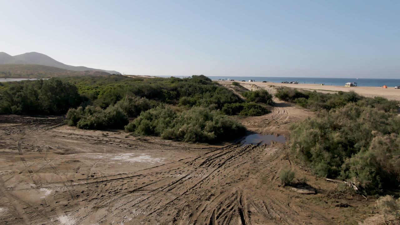 Sandy dirt tracks, coastal scrub, beach, ocean, and hills under a clear sky.