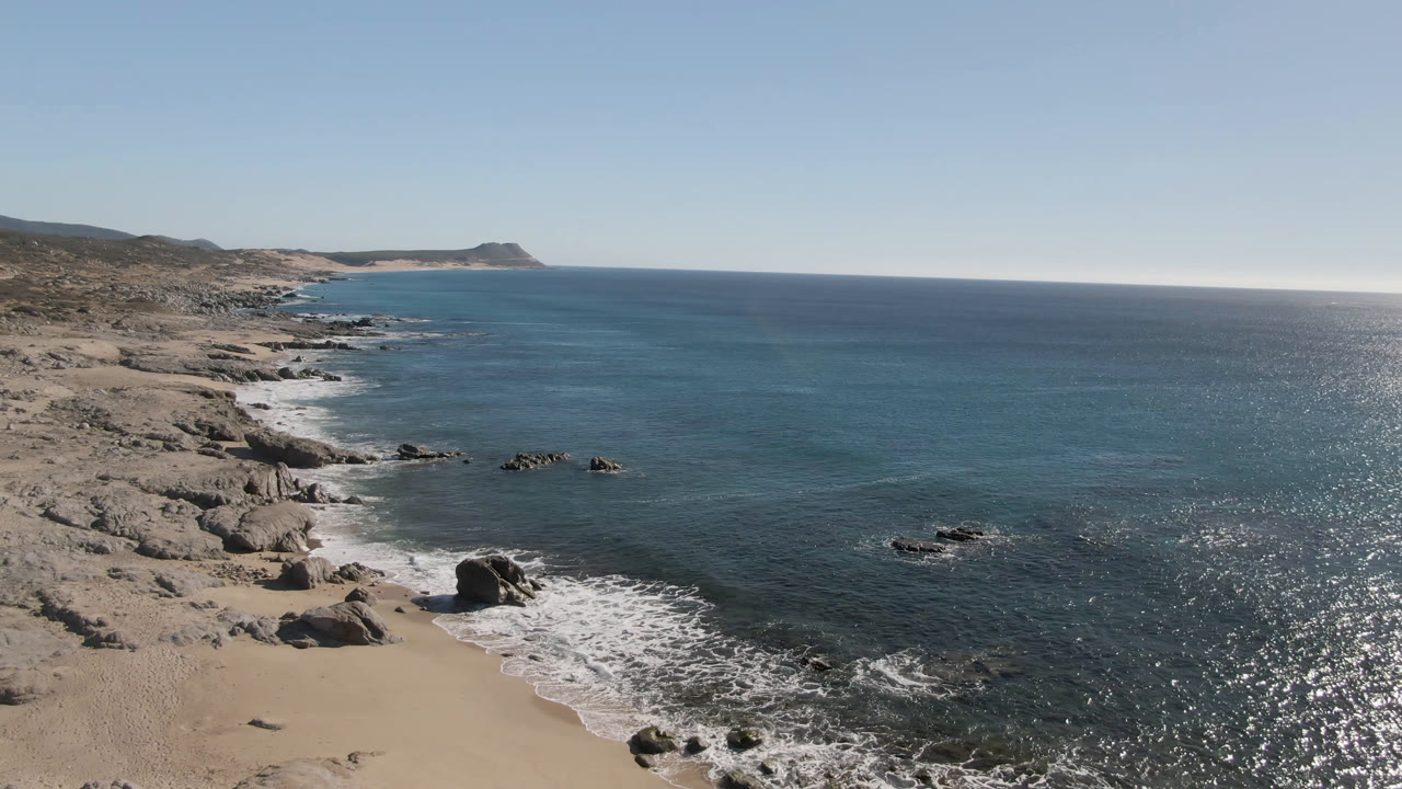 Laguna Beach Los Cabos shoreline seen from above, with a sandy cove, scattered rocks, white wash from small waves, bright water reflections, and no visible people.