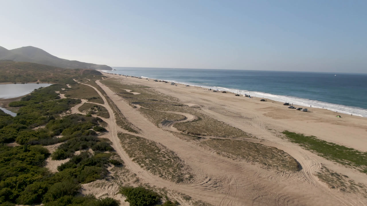 A high aerial frame over Migrino, Baja California Sur shows a broad sandy beach between scrub-covered dunes and blue ocean, with a small water area at left and no visible people.