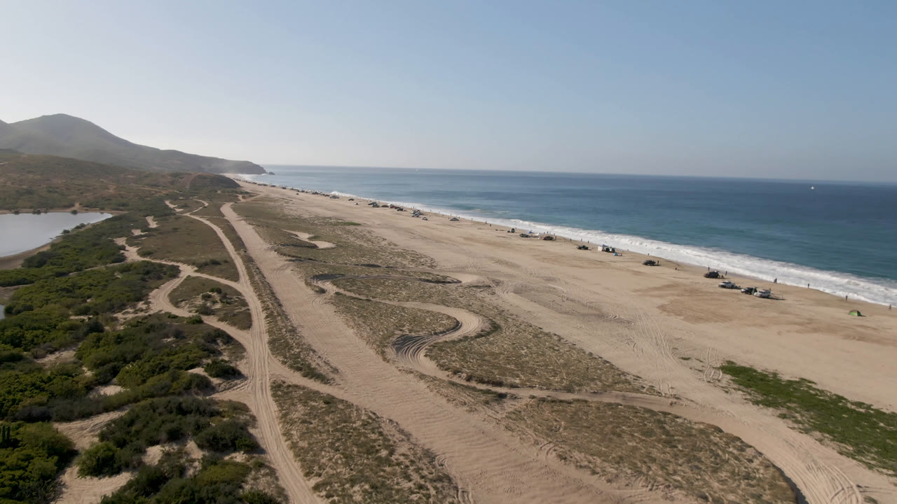 At 0:01, the aerial view over Migrino beach keeps the surf line on the right and brings the curving sandy track pattern nearer the center, with no visible people.