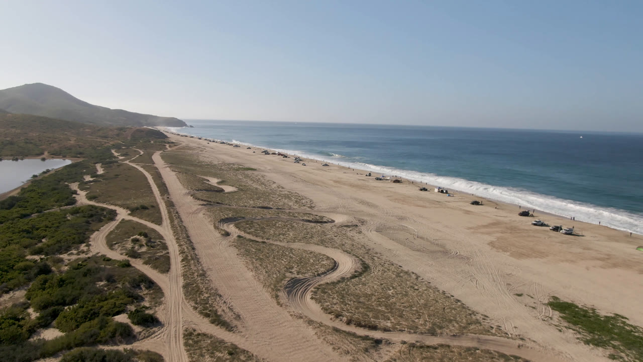 Aerial shoreline with ocean, open sand, dune paths, and distant vehicles.