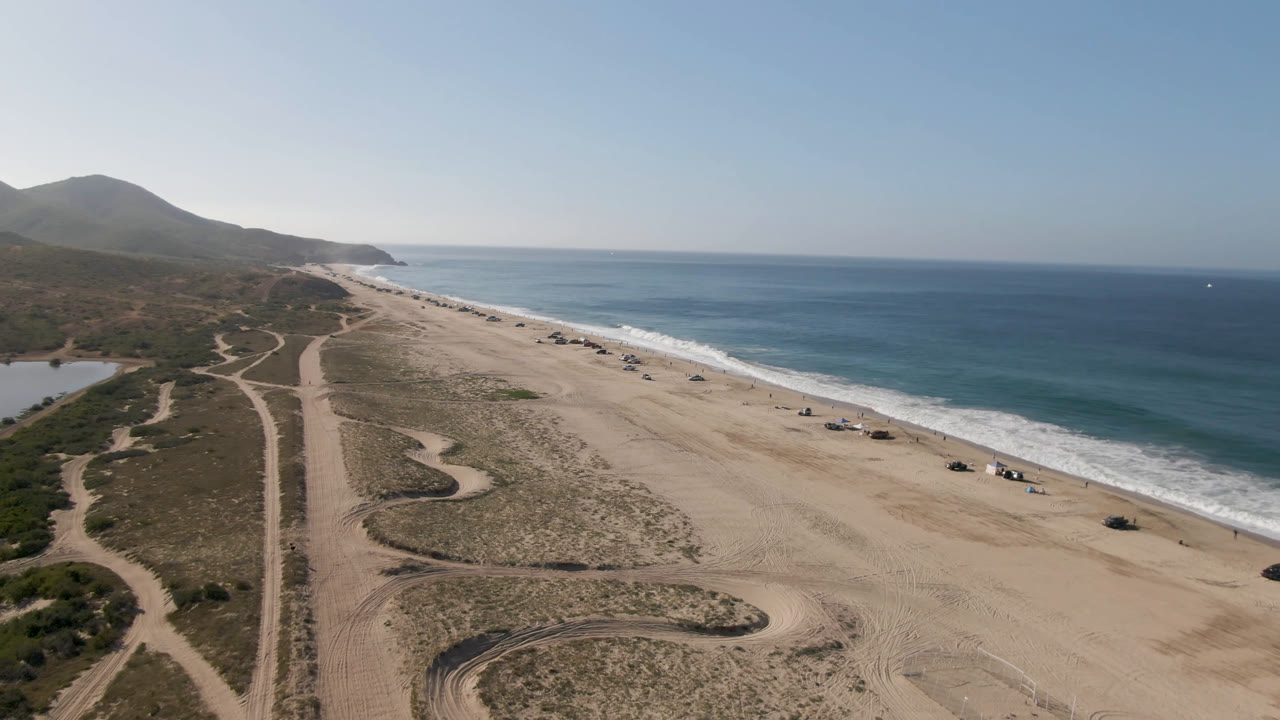 Aerial view of sandy beach, surf, dirt tracks, low hills, and ocean horizon.