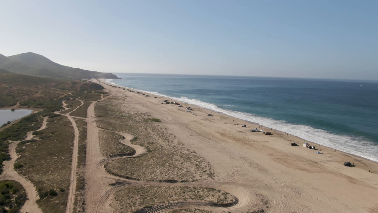 At 0:01, the drone view over Migrino beach centers the sandy access tracks and dune scrub, with parked vehicles near the shoreline and no visible people.
