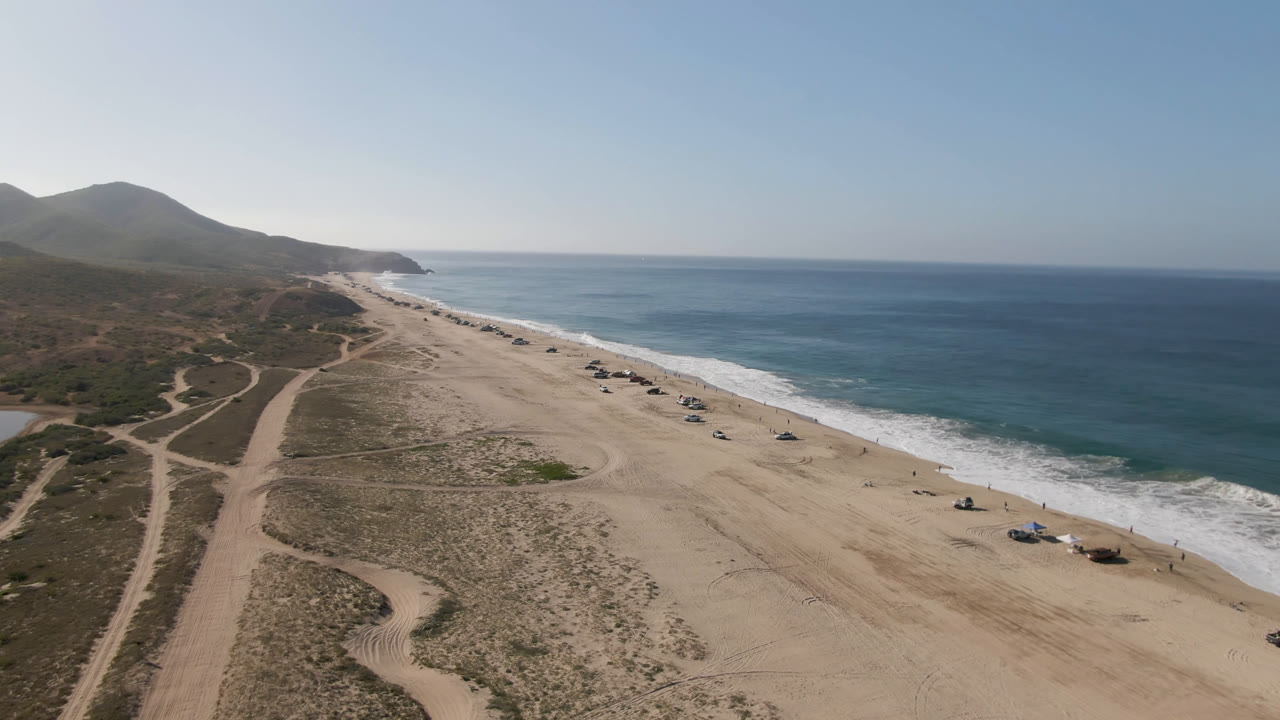 Aerial view over Migrino, Baja California Sur showing dune tracks, a broad sandy beach, blue ocean, distant hills, and no visible people at the opening frame.