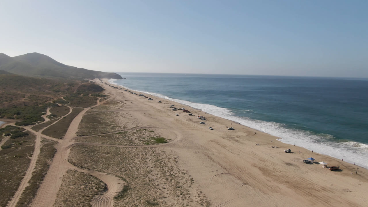 High aerial frame in Migrino, Baja California Sur with beach tracks in the lower frame, scattered vehicles near the waterline, white surf, open ocean, and no visible people.