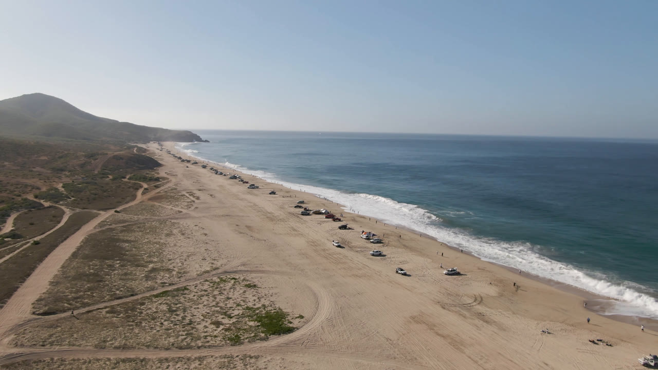 Wide aerial view of Migrino, Baja California Sur, with a sandy beach, parked cars near the waterline, scrub dunes, distant hills, and open blue ocean at the start of the clip.