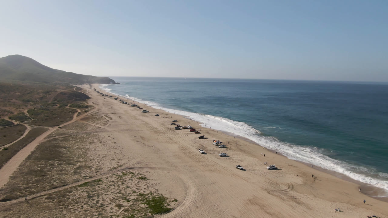 Aerial frame over Migrino beach showing a diagonal shoreline, rows of parked vehicles on pale sand, low coastal vegetation, hills to the left, and a broad ocean horizon.