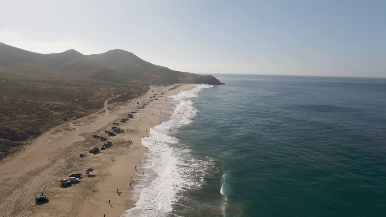 Aerial view of cars parked on a sandy beach beside breaking surf.