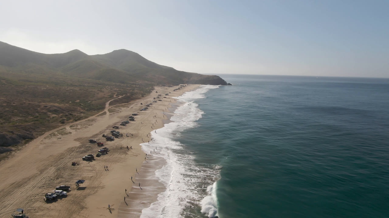 Aerial view over Migrino beach with more of the vehicle line visible on the sand, rolling white water at midframe, open sea on the right, and low hills behind the beach.
