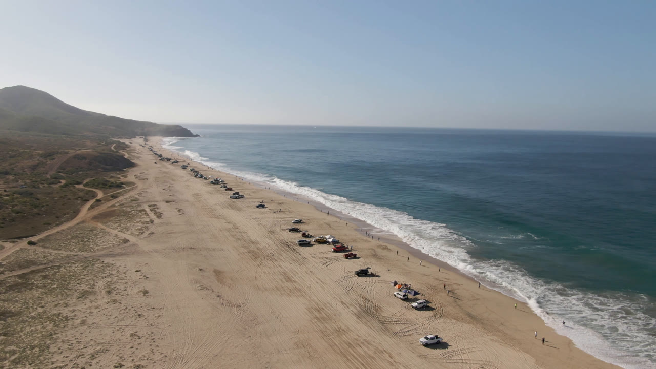 Aerial view of Migrino, Baja California Sur, Mexico, with vehicles parked along a sandy beach, blue ocean to the right, low hills to the left, and no visible people.