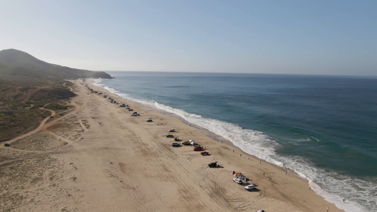 Wide drone frame over Migrino beach showing a diagonal line of parked vehicles on sand, white waves along the shore, open water, and no visible people.