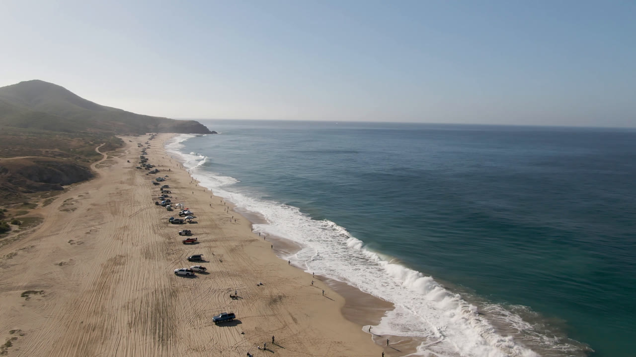 A high aerial frame over Migrino, Baja California Sur shows vehicles lined on the sand, white surf along the right edge, low hills beyond the beach, and no visible people.
