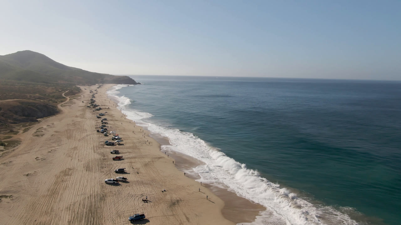 The aerial view in Migrino, Baja California Sur places a long row of parked vehicles down the sandy shore, with ocean water to the right and a hazy headland in the distance.