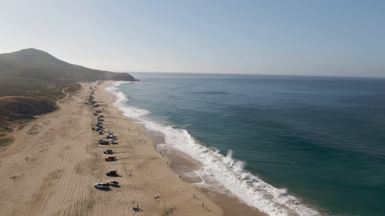Drone perspective over a sandy coastline with vehicles parked near breaking waves.