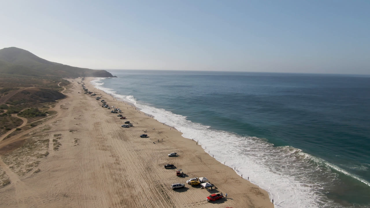 Drone view over Migrino, Baja California Sur showing a long sandy beach with parked cars near the lower frame, white surf to the right, low scrubland to the left, and no discernible people.