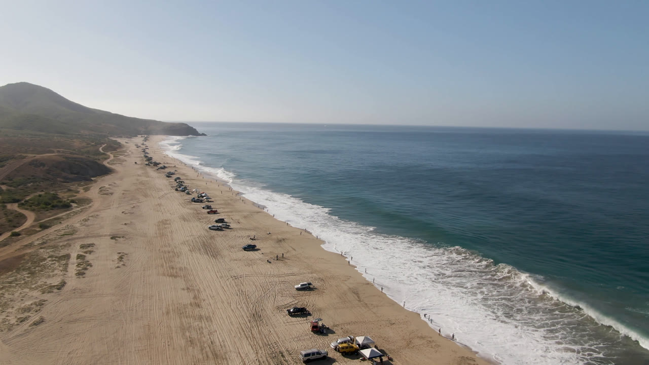 High aerial frame of Migrino beach with a longer line of parked vehicles visible down the sand, surf rolling in from the right, inland dirt paths to the left, and no discernible people.