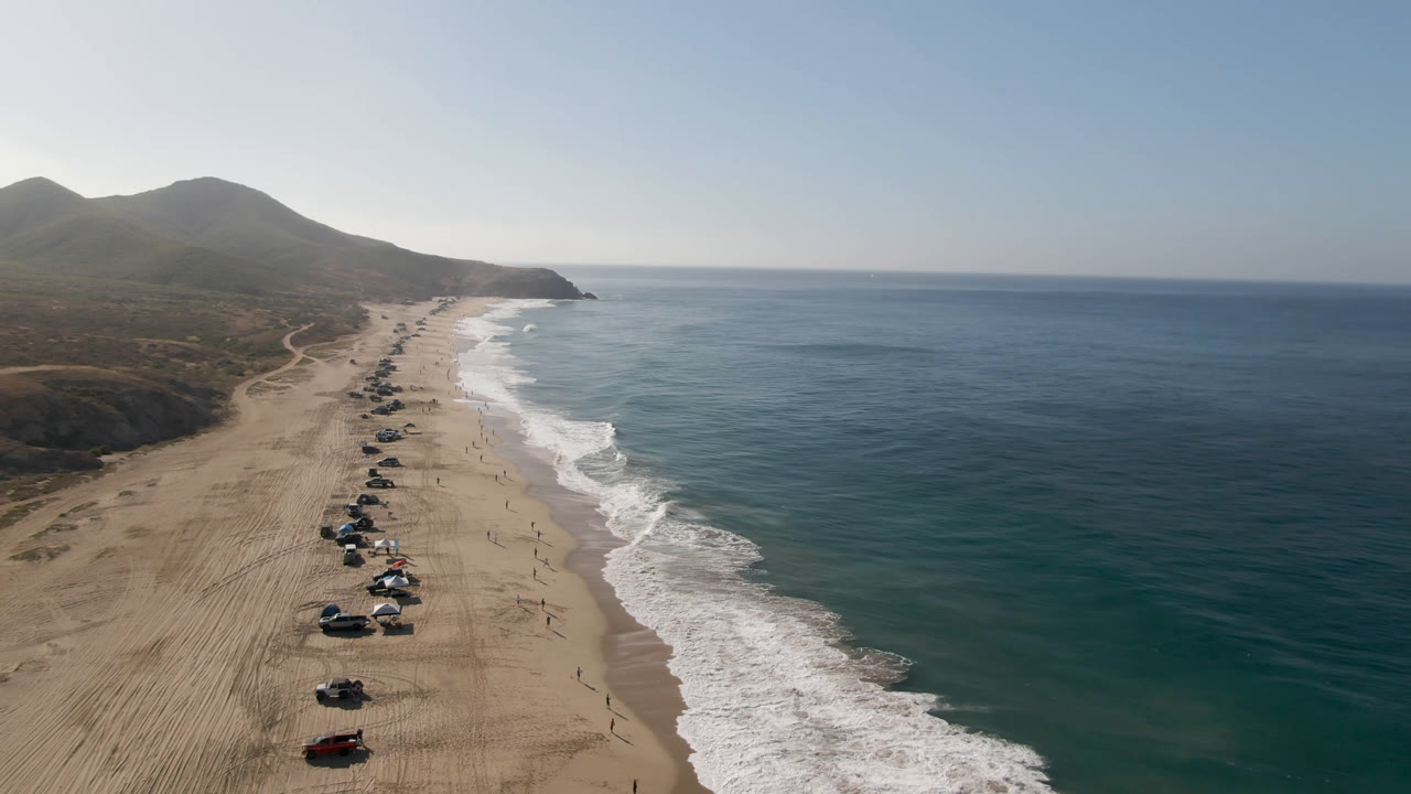 Aerial view of a sandy beach with parked vehicles, surf, ocean, hills, and clear sky.