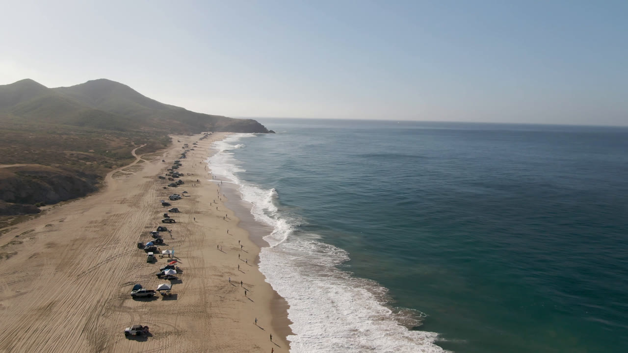 Aerial view over Arafly Beach with a slightly closer line of vehicles and shade setups on the sand, waves curling along the shore, open water to the right, and tiny beachgoers visible.