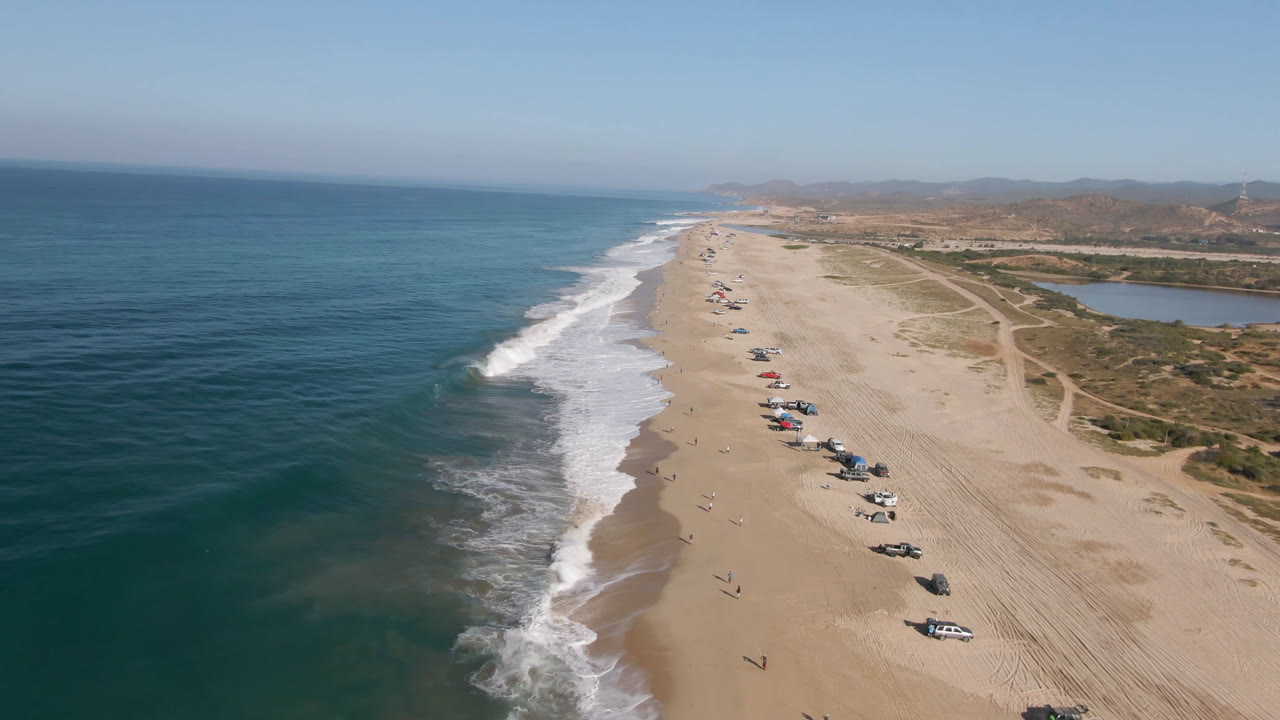 Aerial view over Migrino, Baja California Sur, with blue ocean on the left, white surf along a long sandy beach, parked vehicles on the sand, and distant beachgoers visible.