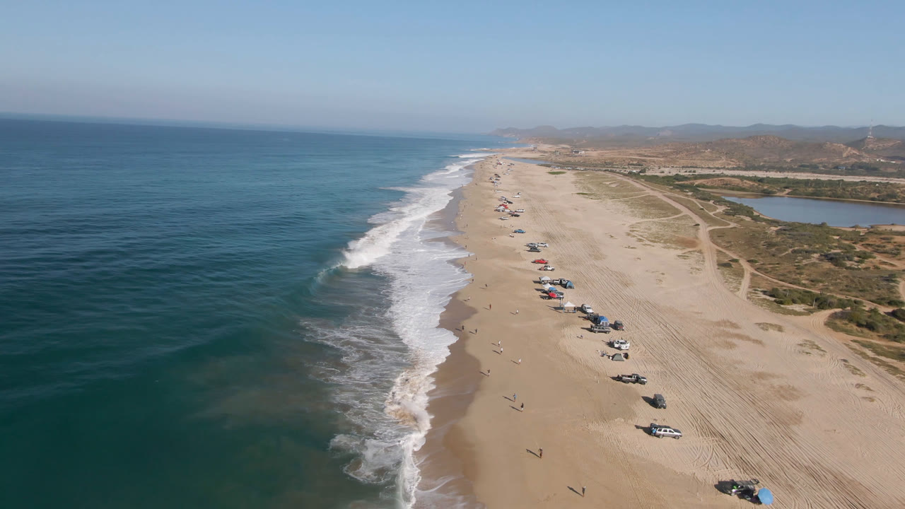 High aerial frame over Migrino beach showing parallel wave foam, a line of parked vehicles on pale sand, scrub-covered dunes, inland water, distant hills, and tiny people on the shoreline.
