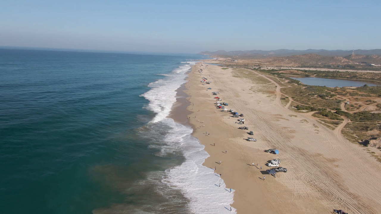 Aerial view of a long beach with ocean waves and parked cars on sand.