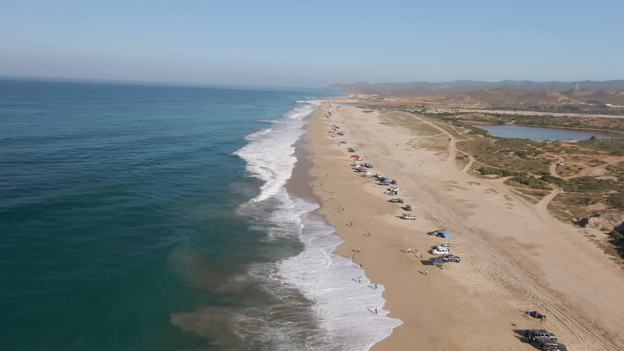 Drone view along Arafly Beach in Migrino, Baja California Sur, with the surf line slightly lower in frame, parked cars spaced down the sand, a lagoon-like water area inland, dry tracks, and no visible people.