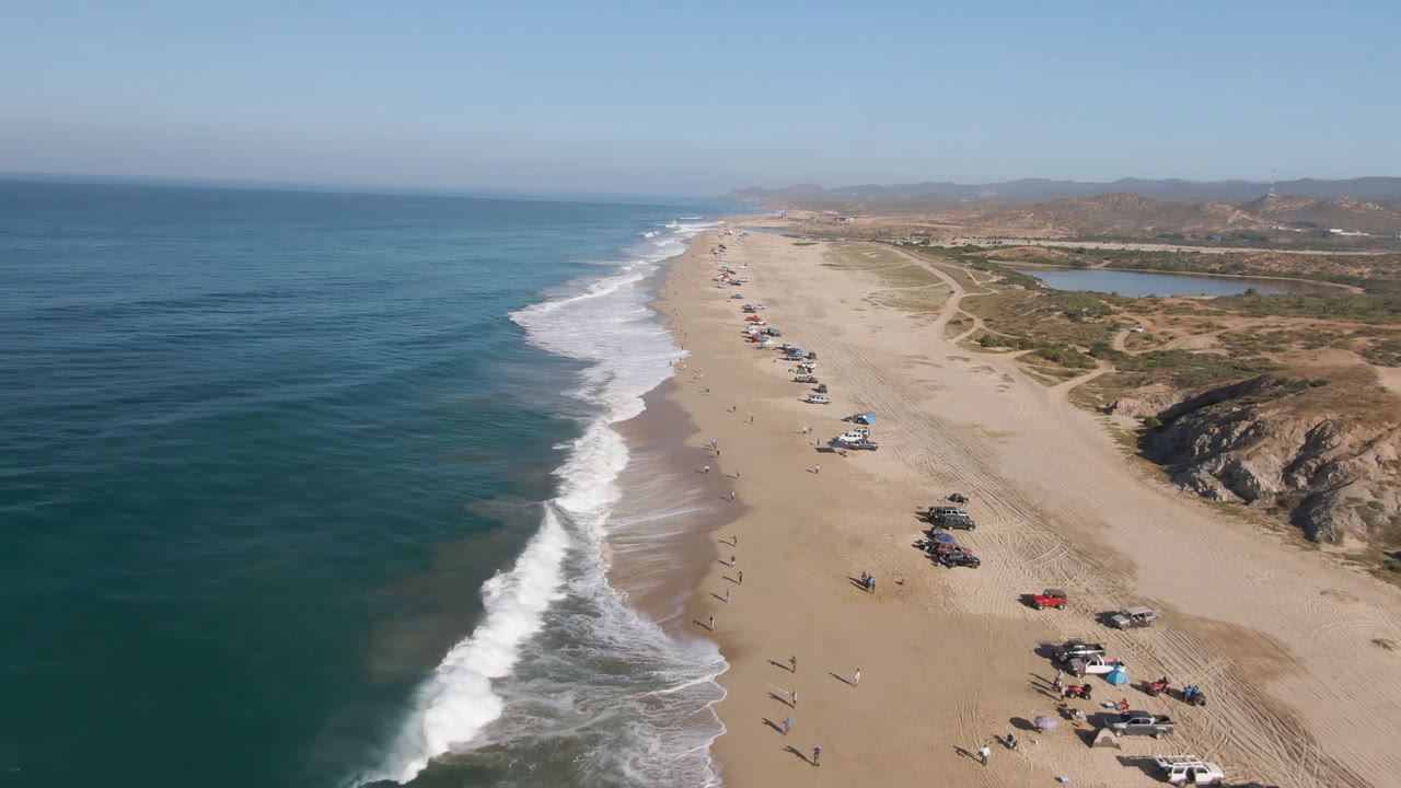 Aerial view of ocean waves beside a long sandy beach with vehicles and small people.