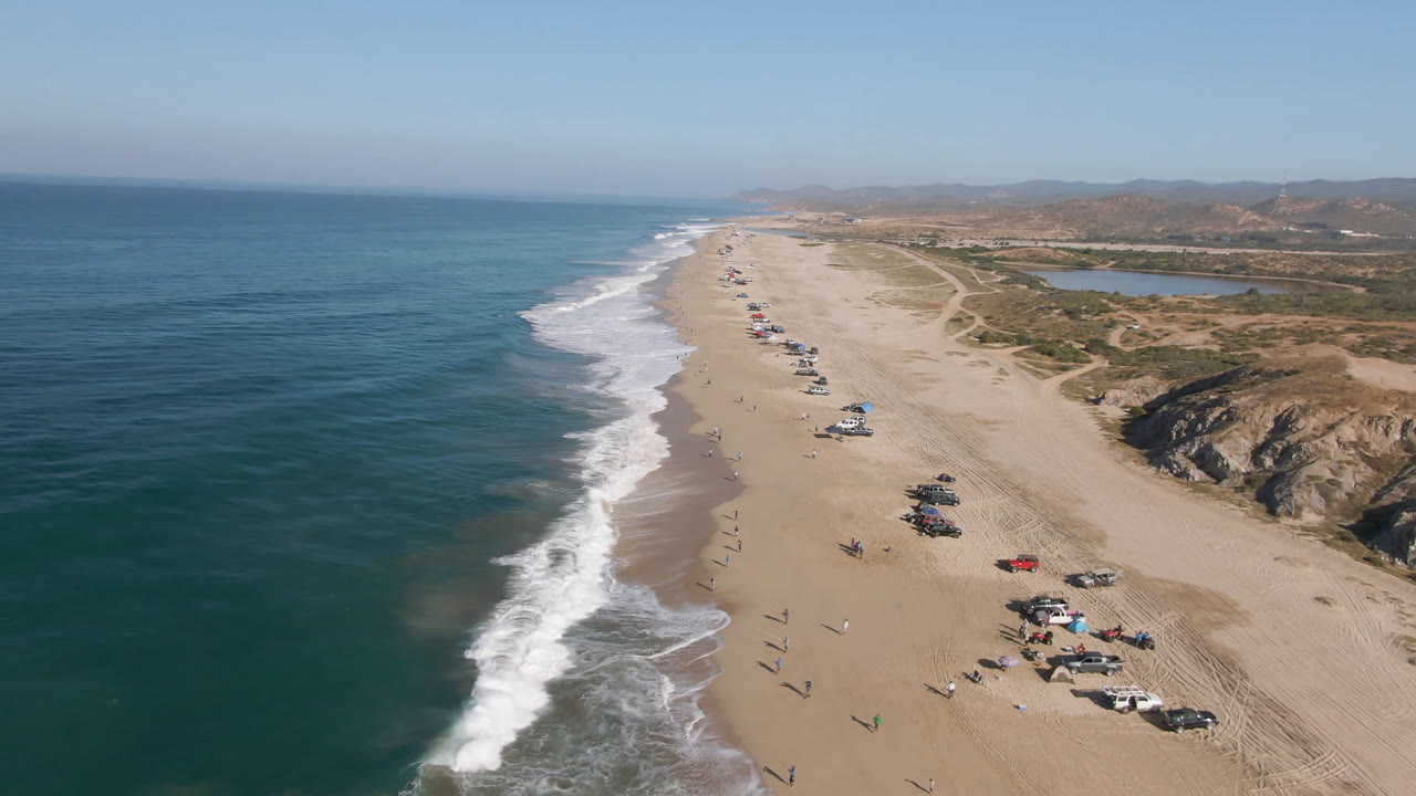 One second into the clip, the drone view shows Arafly Beach with the surf edge slightly farther down frame, clustered vehicles and shade setups on the lower sand, small visible people, and an inland water area beyond the dunes.