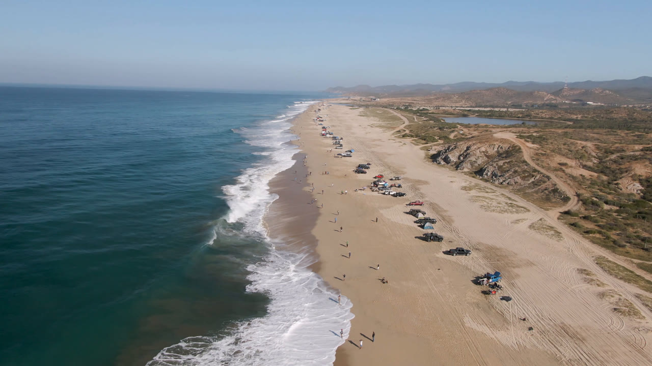 A high aerial frame over Arafly Beach in Migrino, Baja California Sur shows white surf on the left, a long sand strip with parked vehicles, tiny beachgoers, dunes, and hills in the distance.