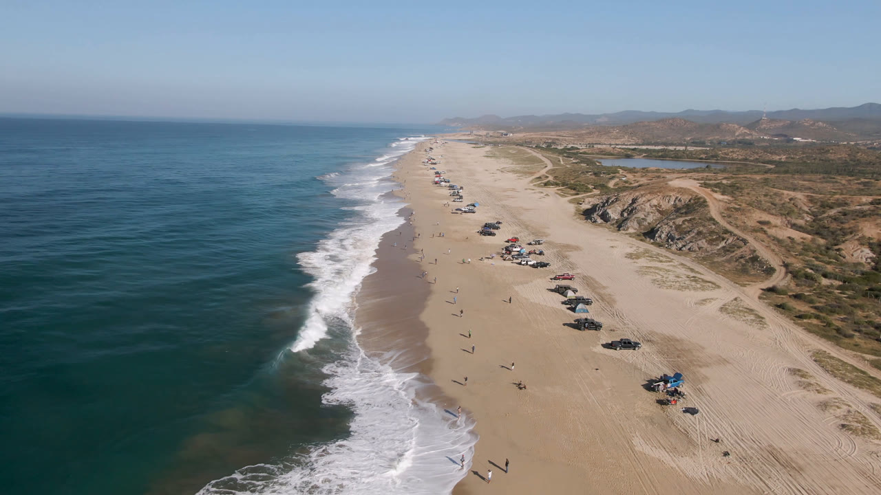 Arafly Beach appears from above with the shoreline running diagonally, white waves breaking along the sand, clustered vehicles near the waterline, small visible people, dune tracks, and arid hills beyond.