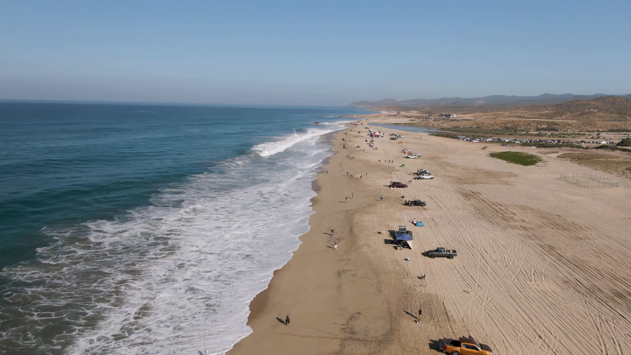 Aerial view of surf along a wide sandy beach with parked vehicles and shade setups.