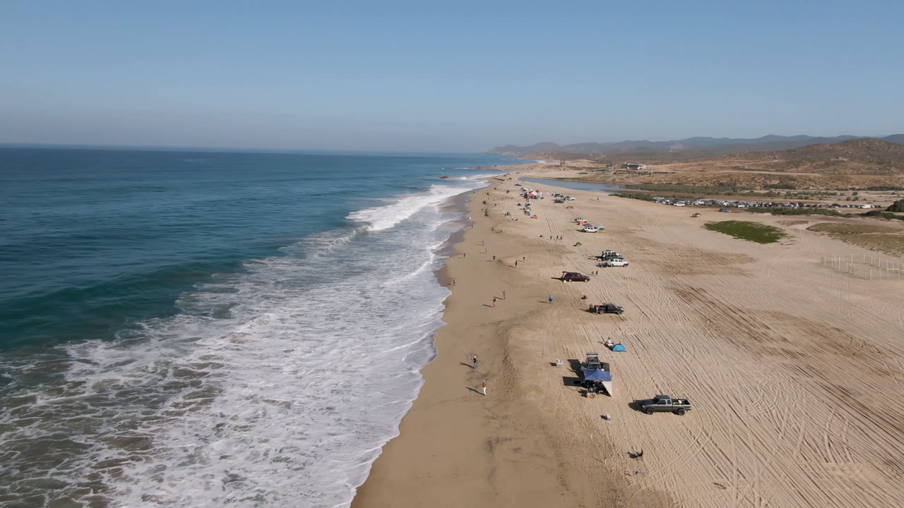 A high drone frame of Arafly Beach in Migrino, Baja California Sur, showing the surf edge curving into the distance, spaced vehicles on the sand, inland scrub, and small distant figures.