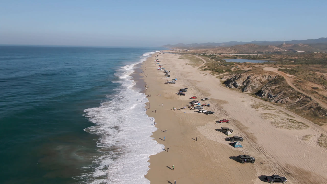Aerial frame over Migrino, Baja California Sur, Mexico showing blue ocean, white surf, a long sandy beach, scattered parked cars, dry hills, and no identifiable people visible.
