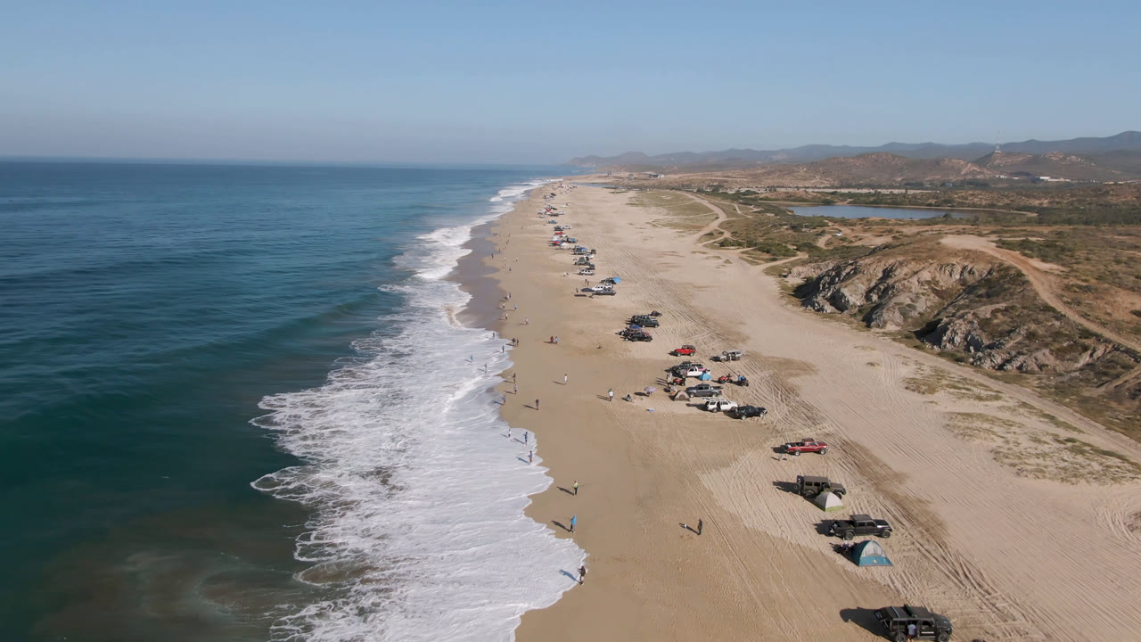 Second aerial frame of the Migrino beach shows the surf pushed farther up the shoreline, vehicles spaced along the sand, inland scrub, low hills, and no identifiable people visible.